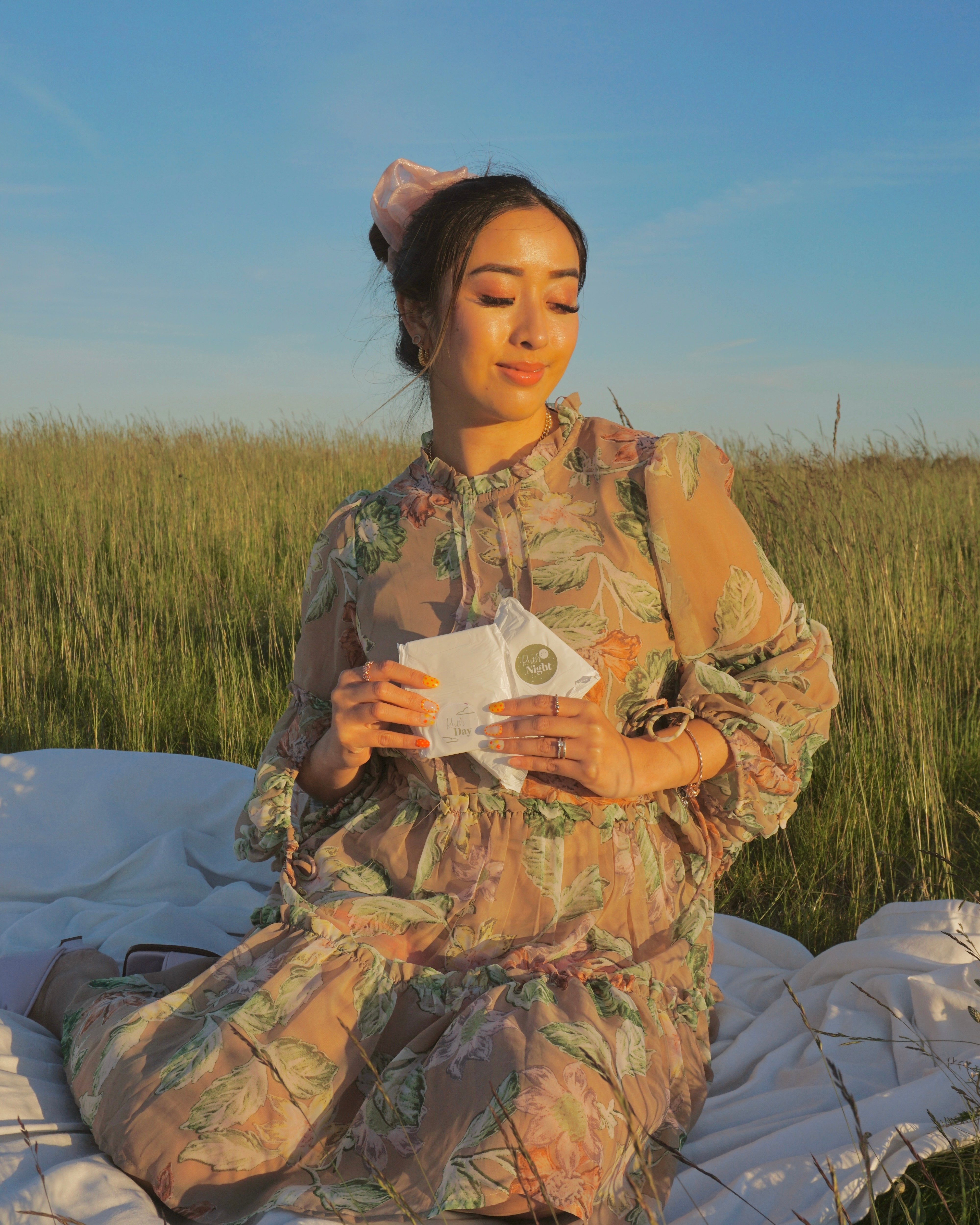 Girl with her eyes closed, sitting in field on a blanket during sunset, holding a Ruth day and a Ruth night pad 